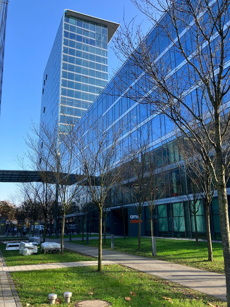 Verglaste Bürogebäude des Skyline Tower München mit durchgehendem Glasdachverbinder; im Vordergrund Grünflächen, junge Bäume und Wegeführung.
