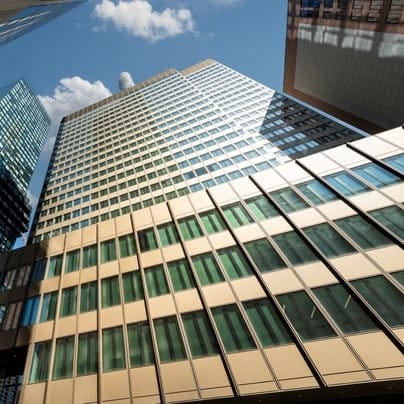 Blick aus der Froschperspektive auf ein modernes Hochhaus mit Glas- und Metallfassade, umgeben von weiteren Hochhäusern unter blauem Himmel.