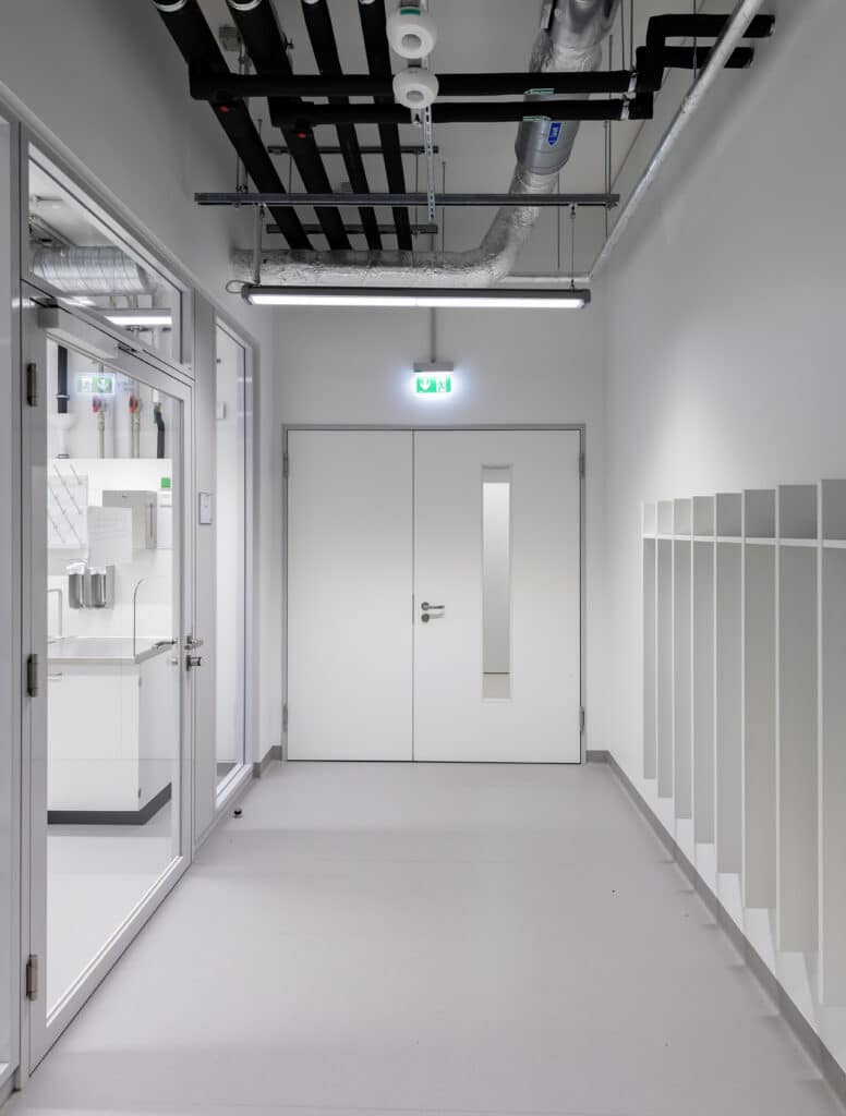 Weißer Technikflur mit sichtbarer Lüftung und doppelten Türen im Klinikbereich.