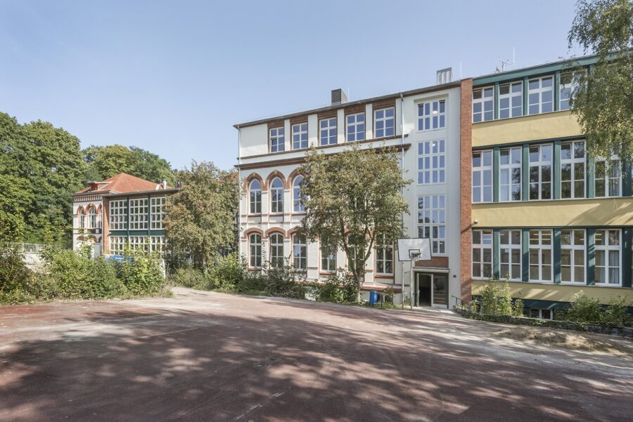 Ansicht eines mehrteiligen Schulgebäudes mit historischer Fassade, großen Rundbogenfenstern und angrenzendem modernen Gebäudeteil, davor ein leerer Schulhof mit Basketballkorb.