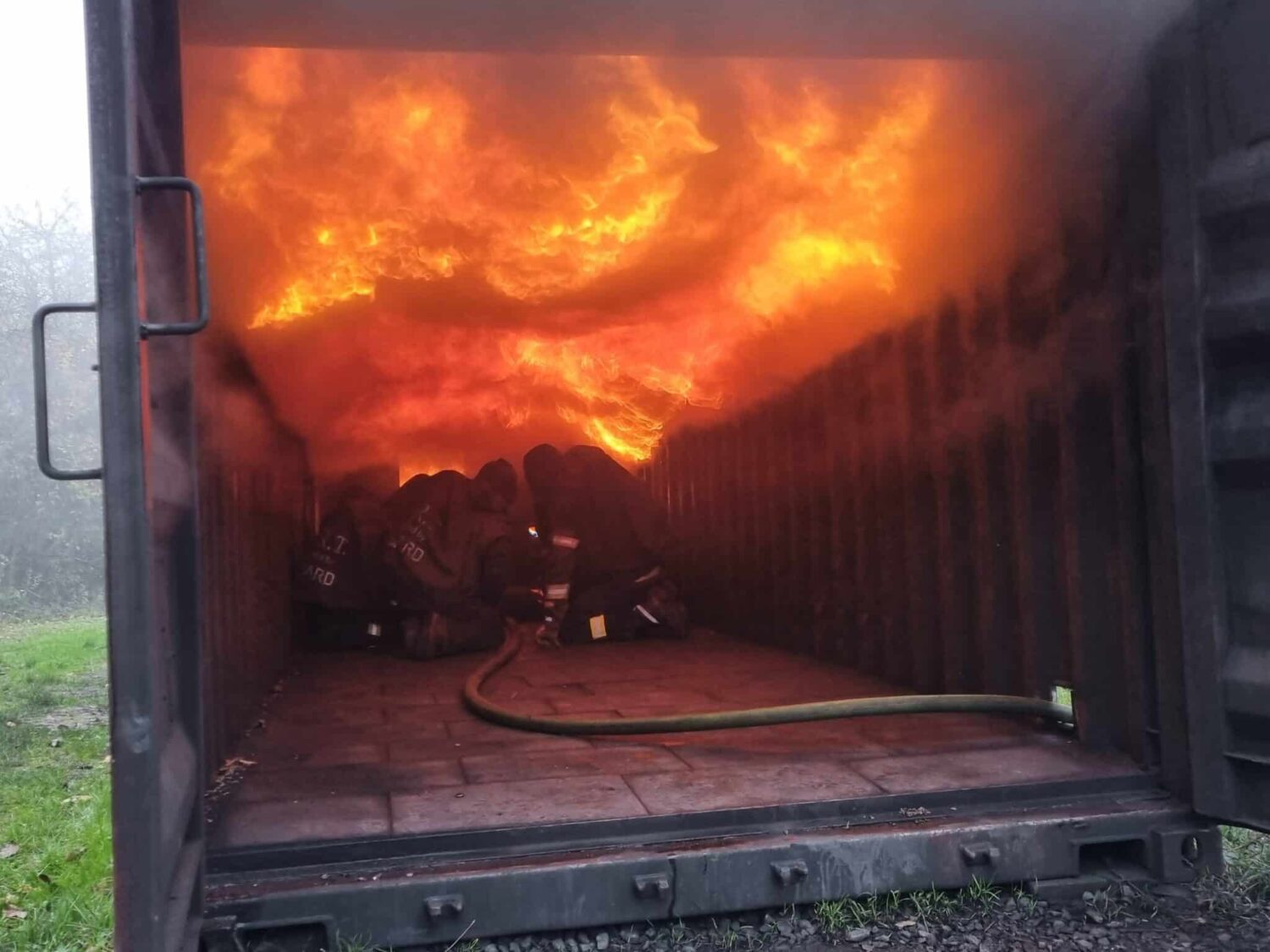 Feuerwehrkräfte knien in einem Brandübungscontainer und bekämpfen einen intensiven Innenbrand; über ihnen breitet sich eine große, flammende Rauchgasdurchzündung aus, die die Decke des Containers vollständig in orangefarbenes Feuer taucht.