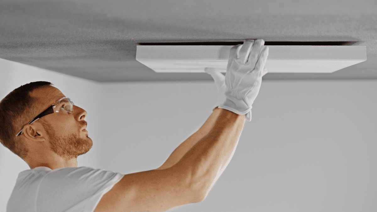 Mann mit Handschuhen und Schutzbrille montiert eine weiße Akustikplatte an einer grauen Decke.