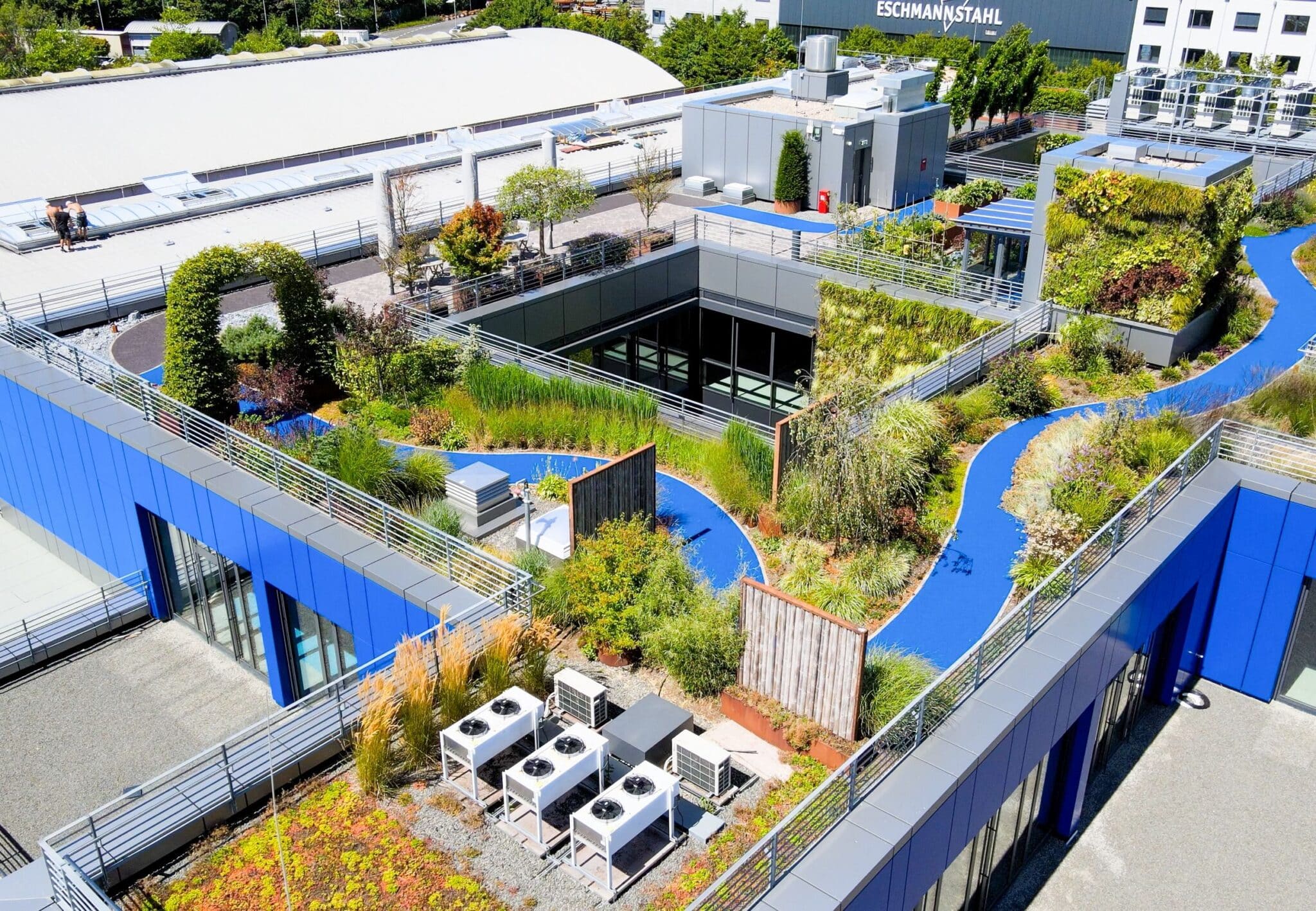 Begrünte Dachlandschaft eines Firmengebäudes mit geschwungenen Wegen, Pflanzbereichen, vertikalen Gärten und technischen Aufbauten in moderner Architektur.