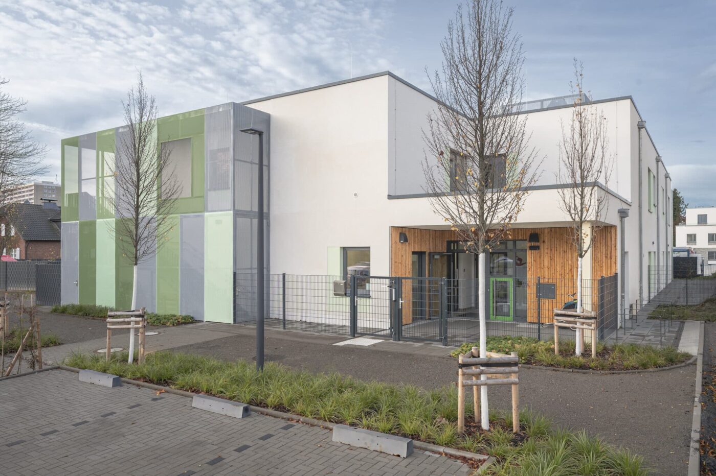 Moderne Kita-Fassade mit hellen Putzflächen, Holzakzenten und farbigen Metallgewebe-Elementen in Grün- und Grautönen; davor neu angelegte Wege, junge Bäume und Eingangsbereich mit Zaun und Tor