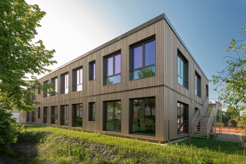 Zweigeschossiger Erweiterungsbau in moderner Holzmodulbauweise mit großen Fenstern und Außentreppe, umgeben von Grünflächen und Bäumen.