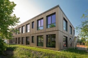Zweigeschossiger Erweiterungsbau in moderner Holzmodulbauweise mit großen Fenstern und Außentreppe, umgeben von Grünflächen und Bäumen.