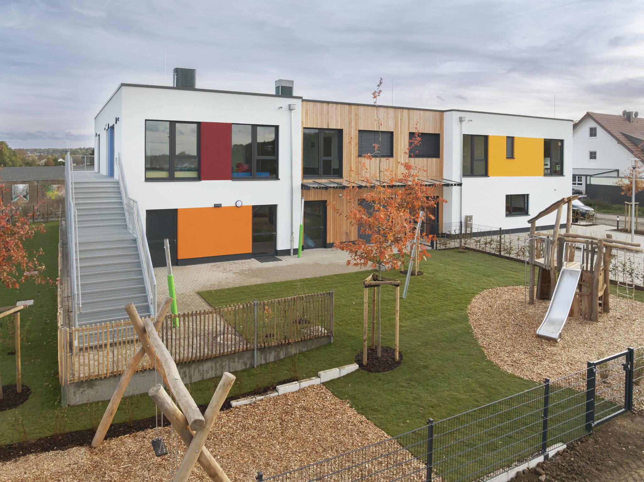 Außenansicht eines modernen zweigeschossigen Kindergartenbaus mit farbigen Fassadenelementen, Holzverkleidung und großem Außenspielbereich.