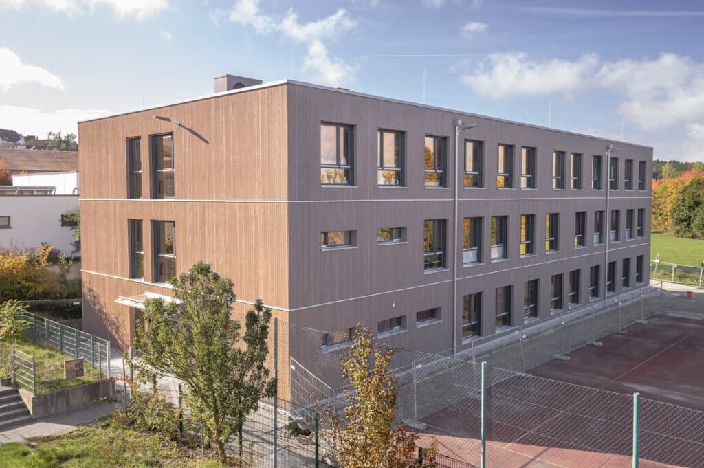 Dreigeschossiges Interims-Schulgebäude in Holzbauweise mit vielen Fenstern, aufgenommen bei sonnigem Wetter; im Vordergrund Bäume und ein umzäunter Außenbereich
