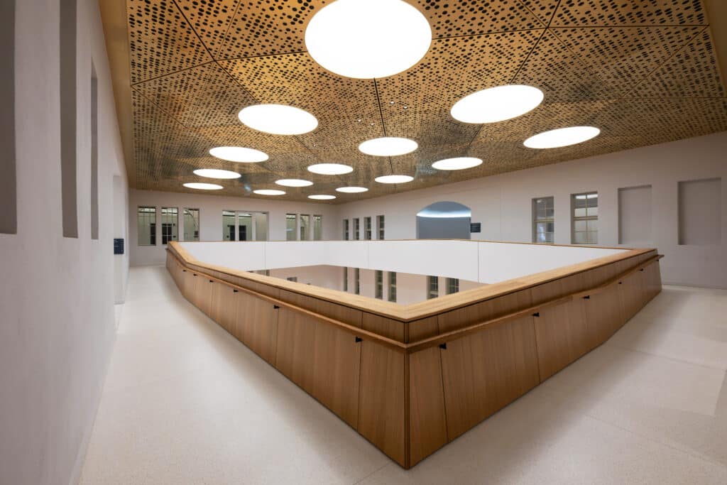 „Innenhof mit umlaufender Holzbrüstung, Decke aus gelochten Holzpaneelen und integrierten Rundleuchten.“
