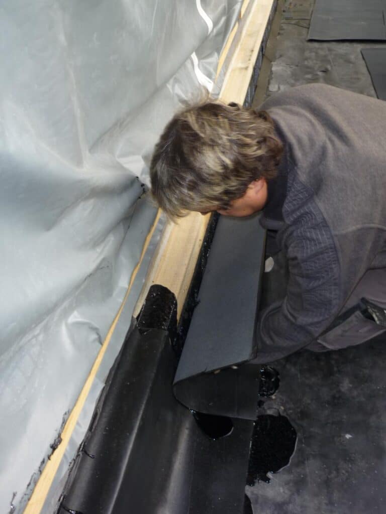 Handwerker bringt eine Bitumenbahn an der aufgehenden Wand eines Flachdachs an.