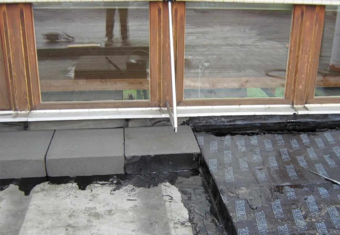 Anschluss einer Flachdachabdichtung an eine Holzfensterfront mit Bitumenbahnen und Dämmung.