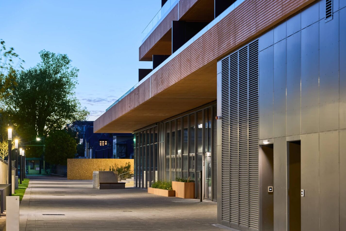 Gebäude mit Metallfassade, Glasflächen und Beleuchtung entlang eines Gehwegs.