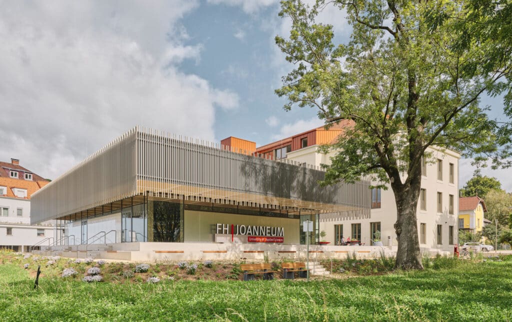 Außenansicht des modernen FH-Joanneum-Gebäudes in Kapfenberg mit umlaufender vertikaler Lamellenfassade und großem begrünten Vorbereich.
