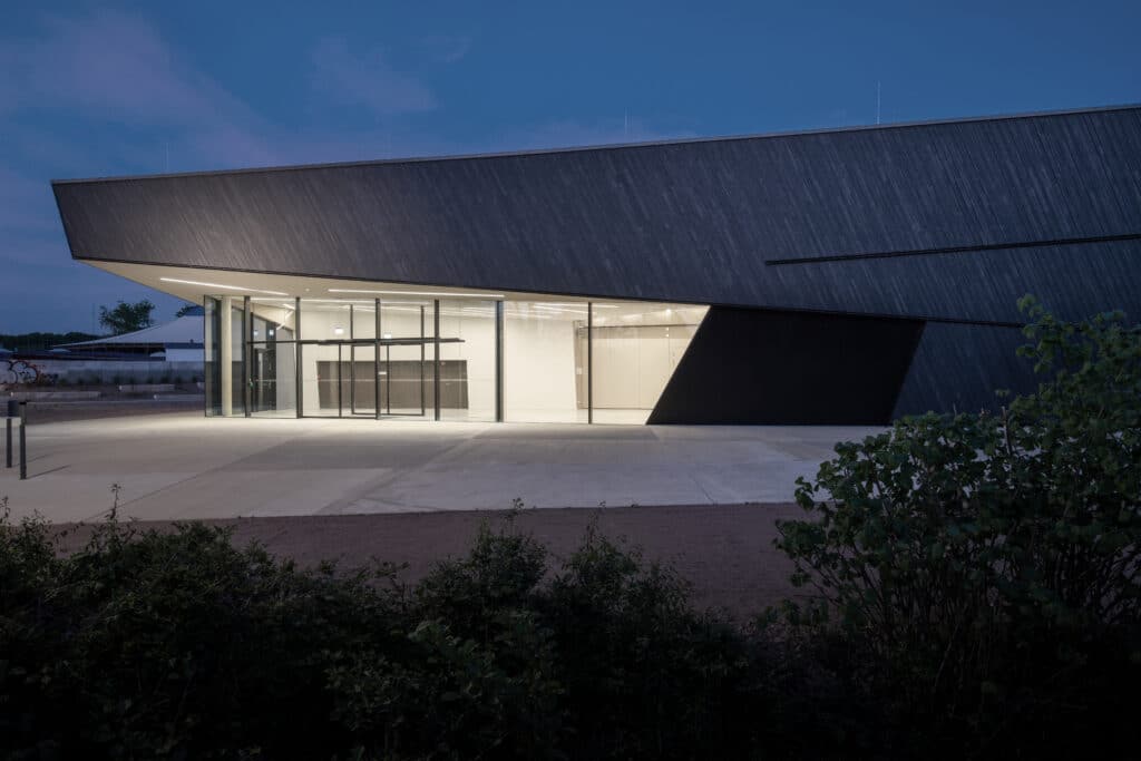 Moderne Veranstaltungshalle in Kuppenheim bei Nacht, beleuchteter Eingangsbereich mit großflächiger Glasfassade und dynamischer Dachform.