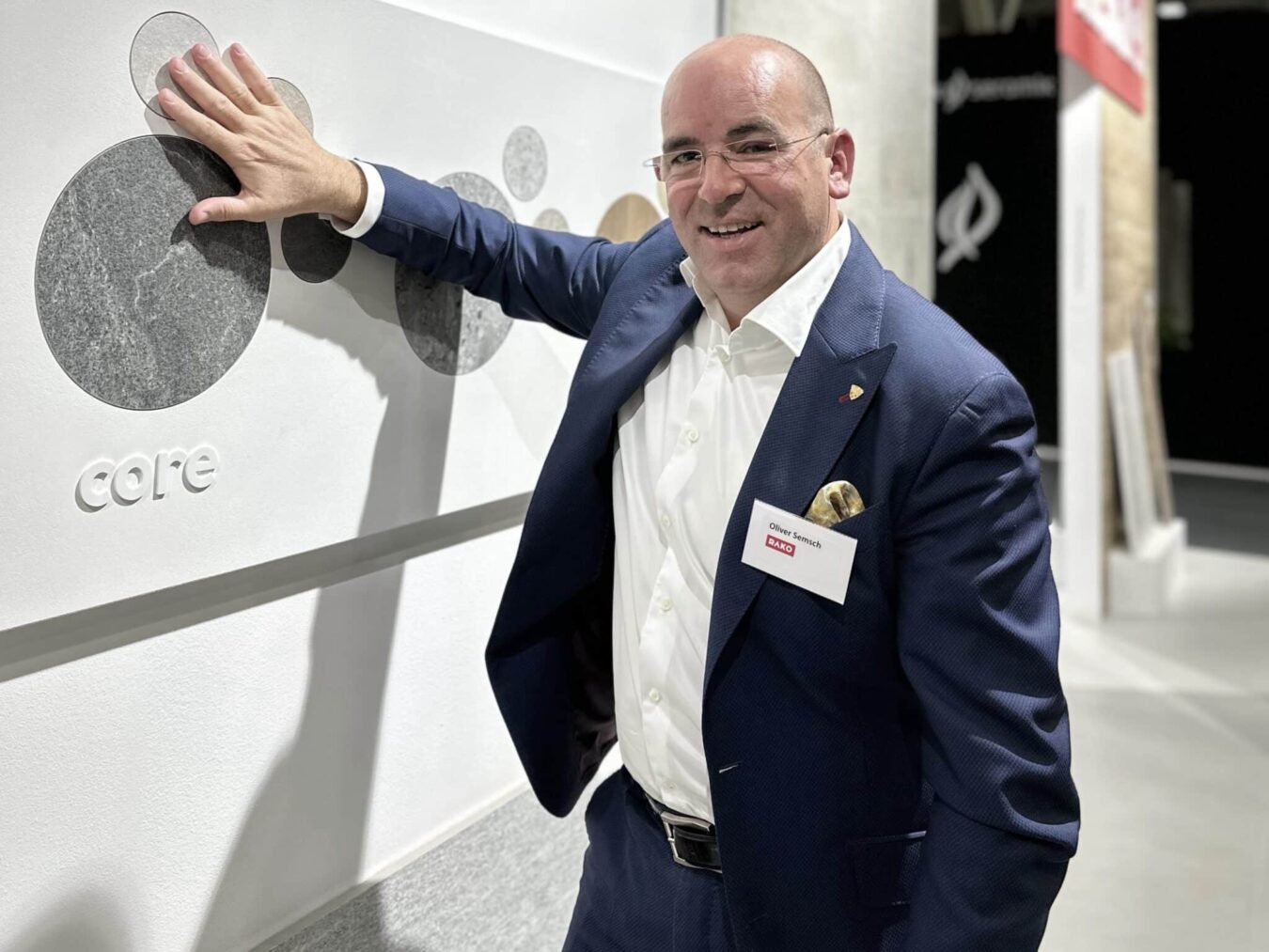 Ein Mann im blauen Anzug mit Namensschild steht an einem Messestand und präsentiert eine Wand mit modernen Oberflächenmustern.