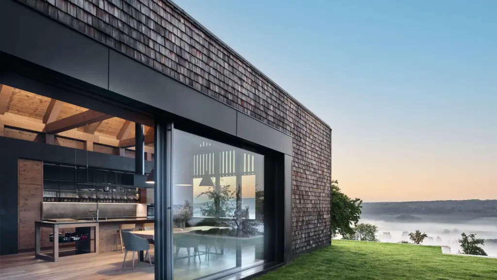 Modernes Wohnhaus mit Holzschindelfassade, großen Glasfronten und Blick über eine neblige Landschaft bei Sonnenaufgang.