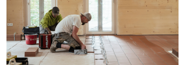 Zwei Handwerker verlegen großformatige Ziegelplatten auf dem Boden eines Wohnraums.