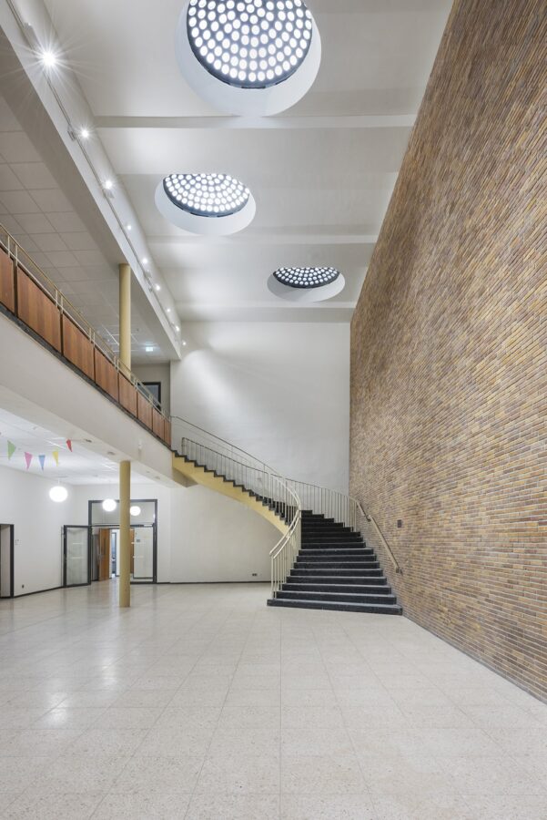 Helles Foyer mit breiter, geschwungener Treppe, hohen Backsteinwänden und runden Oberlichtern, die Lichtpunkte an die Decke projizieren.