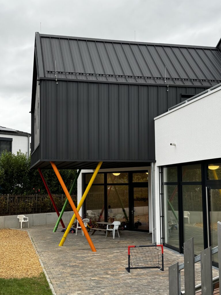 Außenansicht einer Kita mit auskragendem Obergeschoss in dunkler Metallfassade, getragen von farbigen Stahlstützen; überdachter Spielbereich im modernen Kita-Neubau.