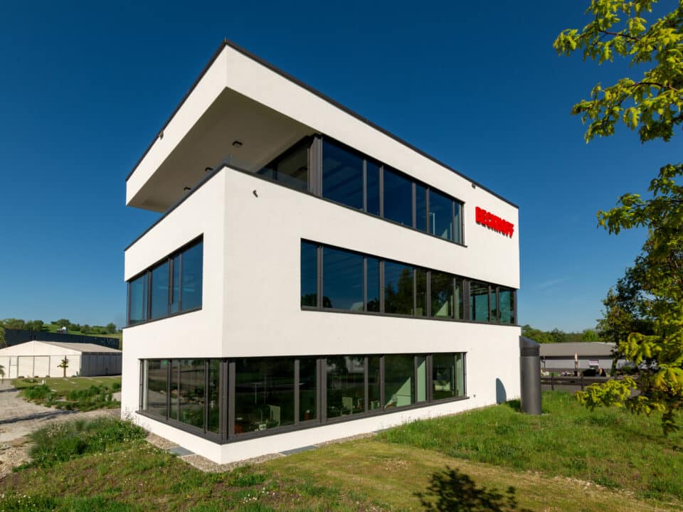 Dreigeschossiges Bürogebäude mit weißer Putzfassade, großen Glasflächen und auskragenden Geschossen; gut sichtbar das rote Firmenlogo an der Fassade, vor klarem blauen Himmel und grüner Umgebung.