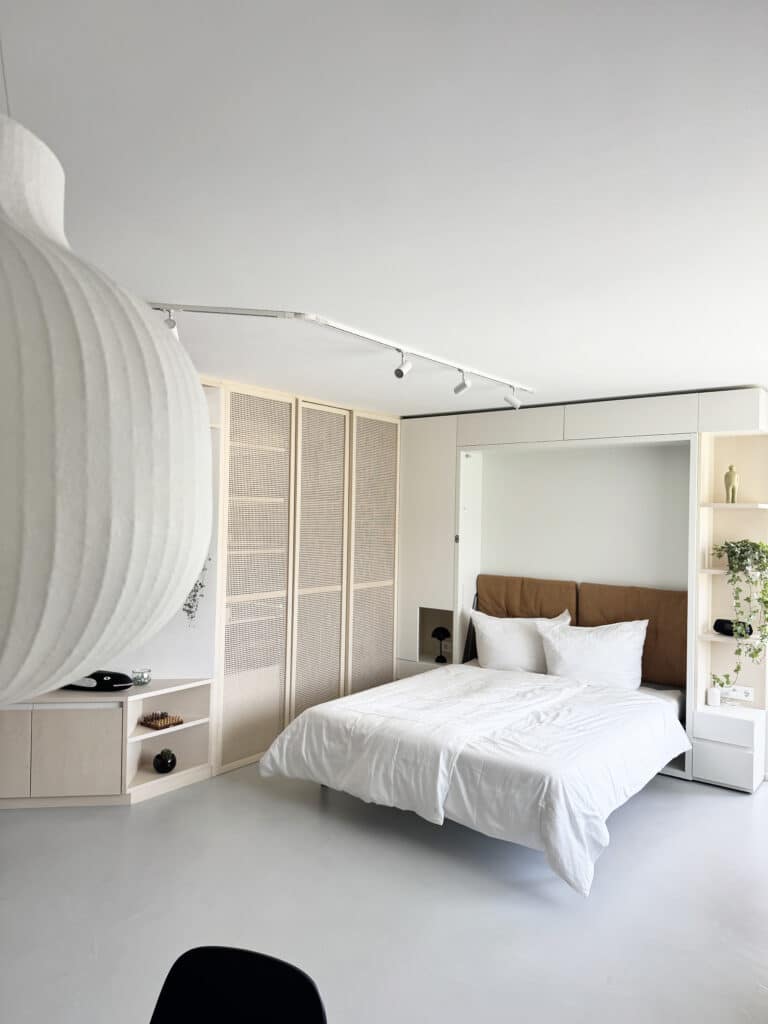 Heller Schlafbereich in einem Studio-Apartment mit Doppelbett, hellen Einbaumöbeln, Schiebetüren mit Geflechtfüllung und offenen Regalen. Minimalistische Einrichtung mit warmen Texturen und natürlichem Licht