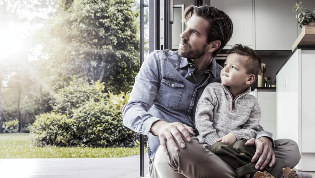 Vater und Sohn sitzen entspannt im offenen Wohnraum vor einer großen Glas-Schiebetür und blicken gemeinsam in den sonnigen Garten.