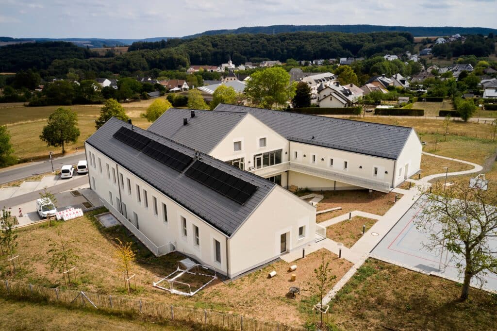 Luftbild eines modernen Gebäudekomplexes mit großflächig integrierten Photovoltaikmodulen auf dem Dach, umgeben von Grünflächen und Wohnbebauung.