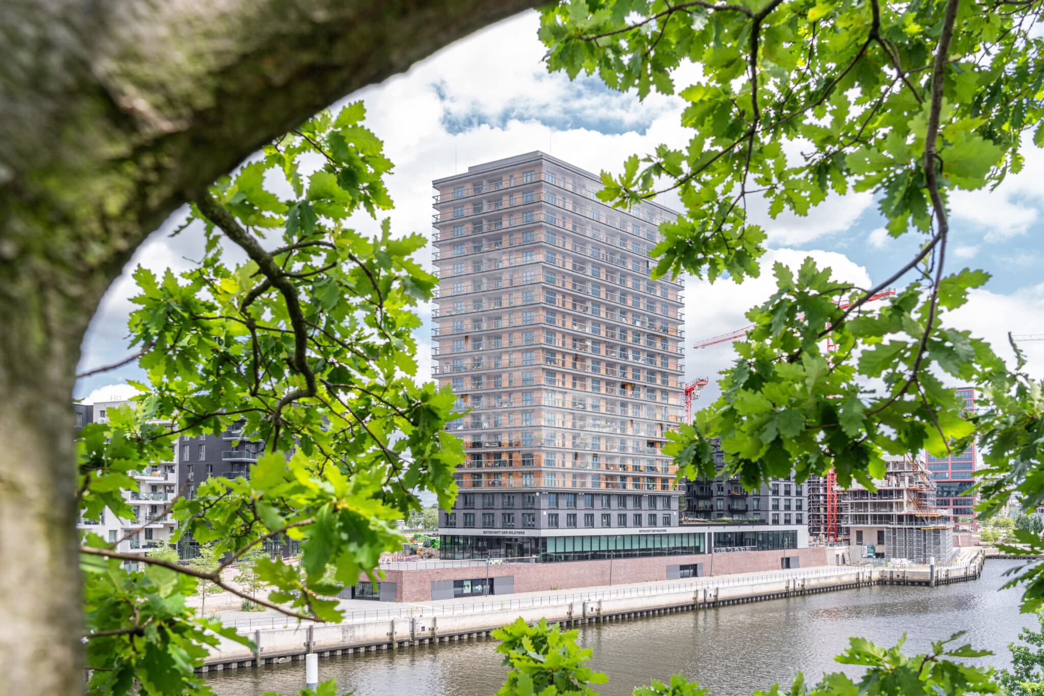 Hochhaus mit gemischter Nutzung am Flussufer, eingerahmt von Bäumen – modernes Stadtquartier im Bau