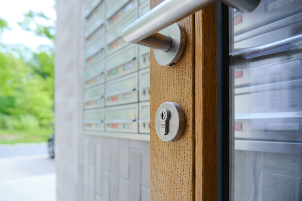 Detailaufnahme einer Holztür mit Edelstahlgriff und Schließzylinder von ASSA ABLOY, im Hintergrund Briefkastenanlage eines Mehrfamilienhauses.
