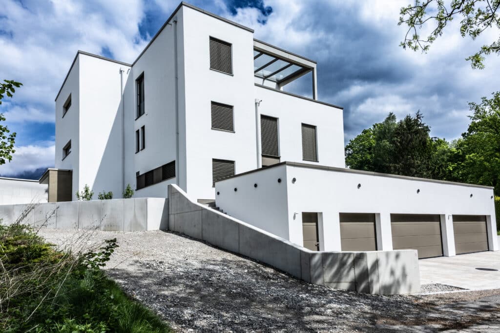 Dreigeschossiges Wohnhaus mit kubischer Formensprache, weißer Putzfassade und integrierter Garagenanlage auf abschüssigem Grundstück.