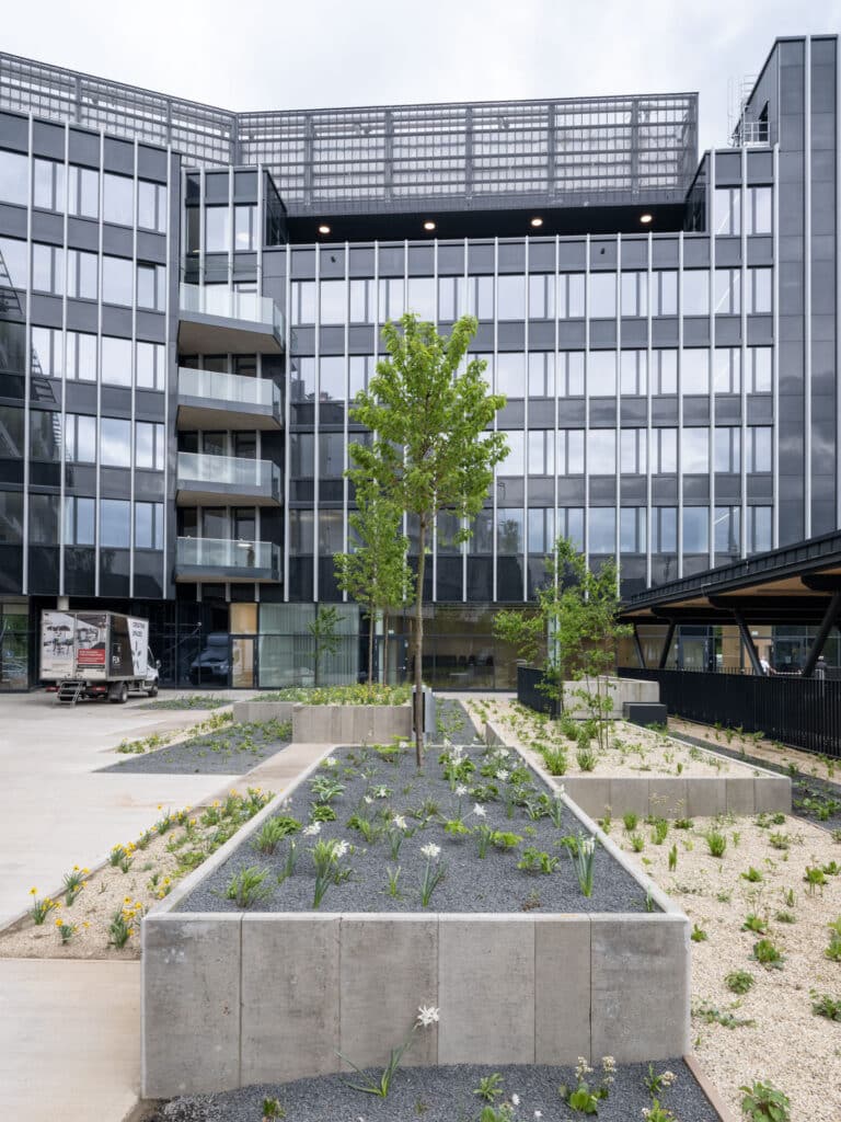 Außenansicht eines modernen Gebäudes mit schwarzer Metallfassade und bepflanztem Innenhof mit Hochbeeten.