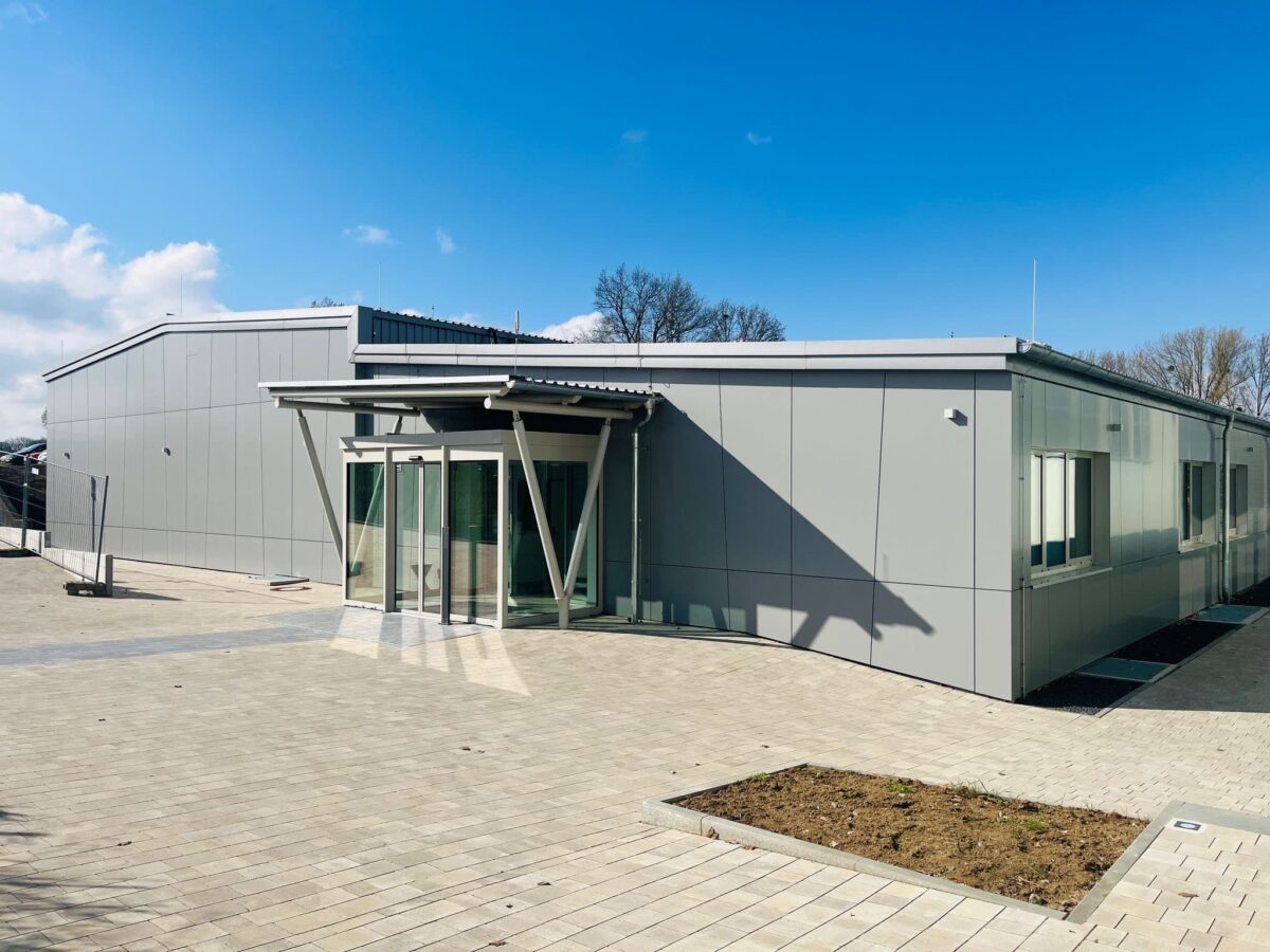 Eingangsbereich eines modernen Hallenbads mit grauen Metallpaneelen, Glasvorbau und großem gepflastertem Vorplatz.