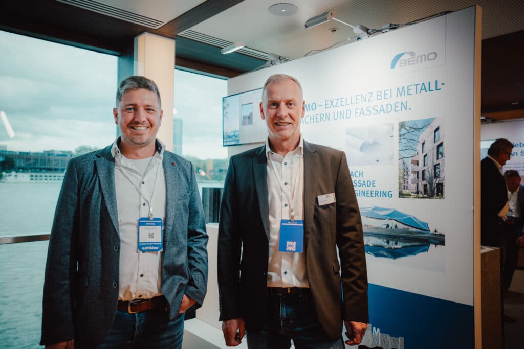 Zwei Mitarbeiter von BEMO präsentieren sich am Messestand auf der ARCHIPOINT Rivercruise 2025 mit Fokus auf Metallfassaden und Dachsysteme.