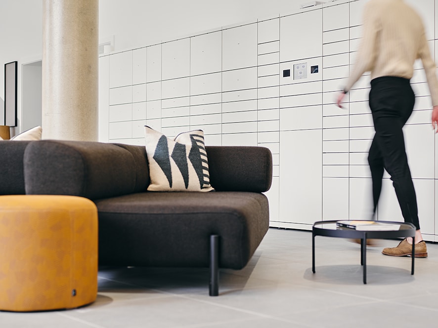 Moderne Lobby mit dunklem Sofa, gelbem Hocker und weißen Paketfächern im Hintergrund, Person geht durch den Raum