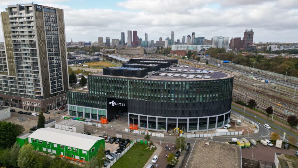 Große Luftaufnahme des modernen Polizei-Neubaus in Den Haag mit gebogener Glasfassade, Dachanlagen und umliegender Stadt- und Bahnstruktur.