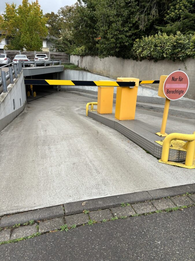 Schranke und Zugangskontrollsystem an der Einfahrt einer Tiefgarage mit geneigter Betonrampe.