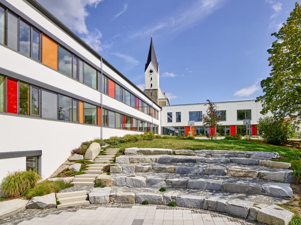 Sanierte Mittelschule Hengersberg mit moderner Fassade, farbigen Fensterelementen und neu gestaltetem Schulhof vor einer Kirche im Hintergrund