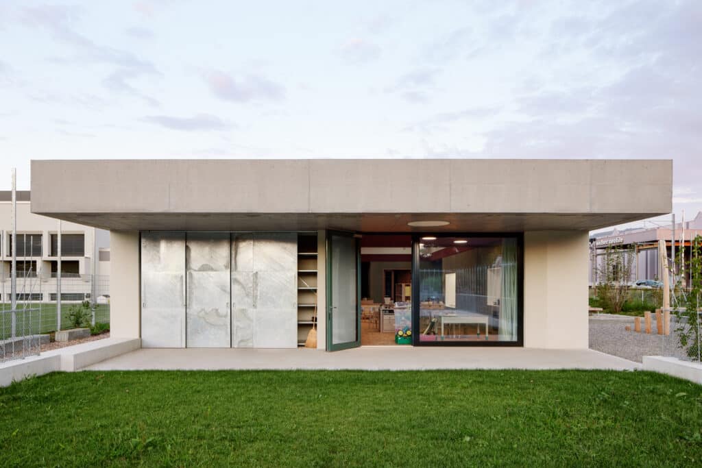 Außenansicht einer modernen Kita mit Betonvordach, großem Schiebetor und bodentiefen Fenstern, Blick auf den Innenraum.