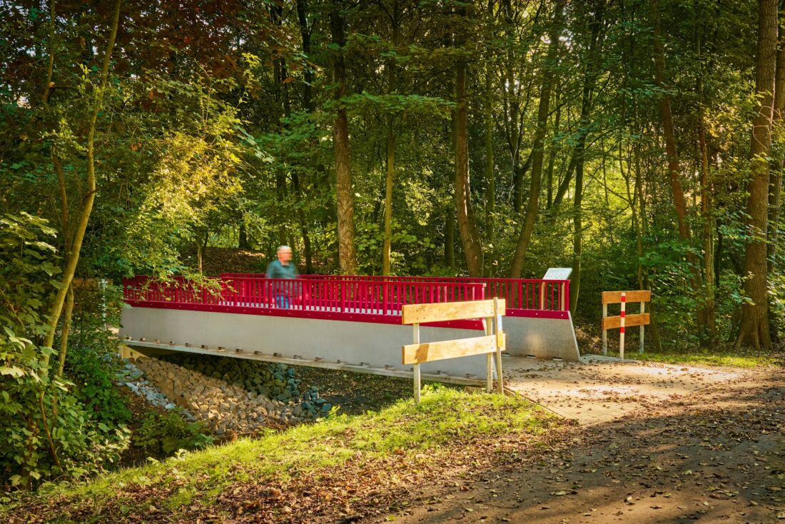 Fußgängerbrücke aus Carbonbeton mit rotem Geländer im Grünen in Oldenburg, aufgenommen im warmen Sonnenlicht eines Waldwegs.