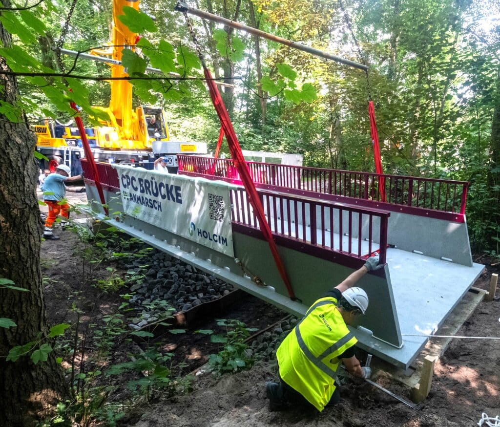 Arbeiter montieren eine CPC-Brücke mit roten Geländern im Waldgebiet von Oldenburg, unterstützt von einem gelben Mobilkran. Die Brücke trägt ein Holcim-Banner mit QR-Code.