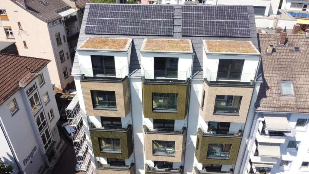 Luftaufnahme eines neu errichteten Stadthauses mit begrünten Flachdächern, Glasbalkonen und großflächiger Photovoltaikanlage.