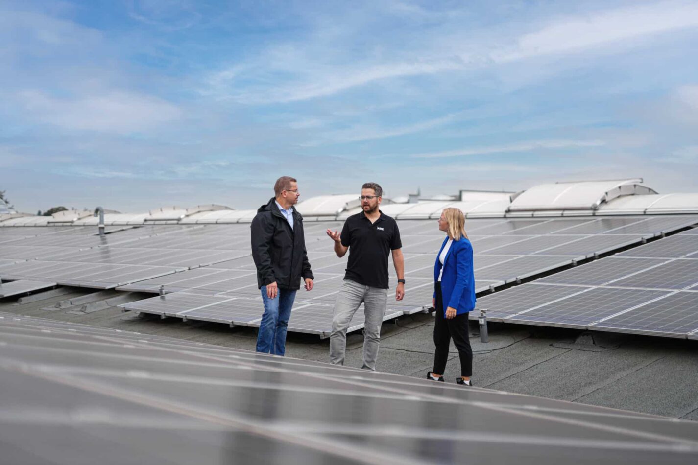 Drei Personen stehen auf einem Hallendach zwischen großen Photovoltaikmodulen und sprechen miteinander.