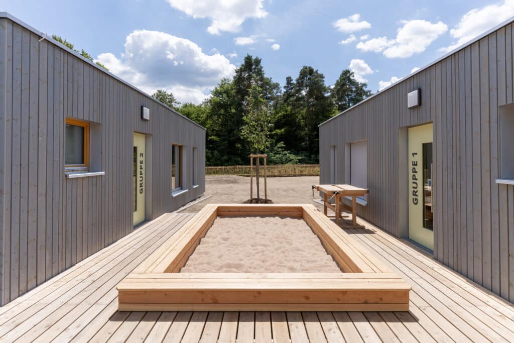 Blick in den Innenhof einer Kita, mit Sandkasten, Holzdeck und den Eingängen zu den Gruppenräumen.