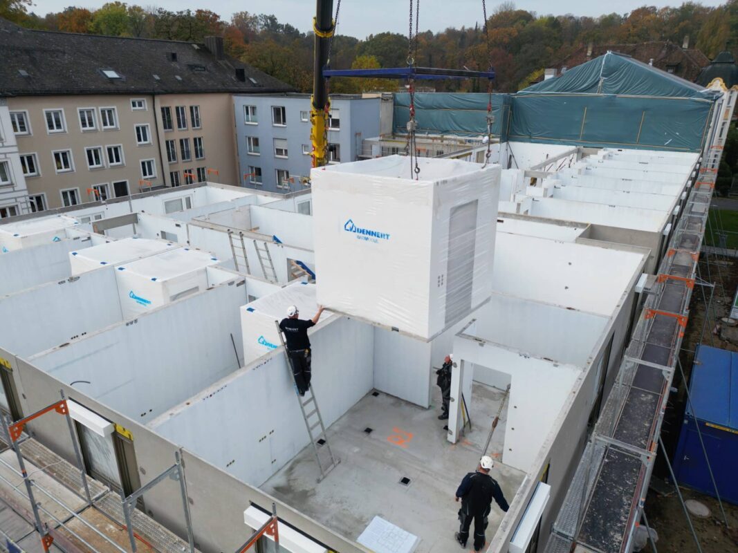 Ein Kran hebt ein vollständig verpacktes Badmodul von Dennert in die Rohbaukonstruktion eines Pflegezentrums ein, während Monteure das Modul präzise positionieren.