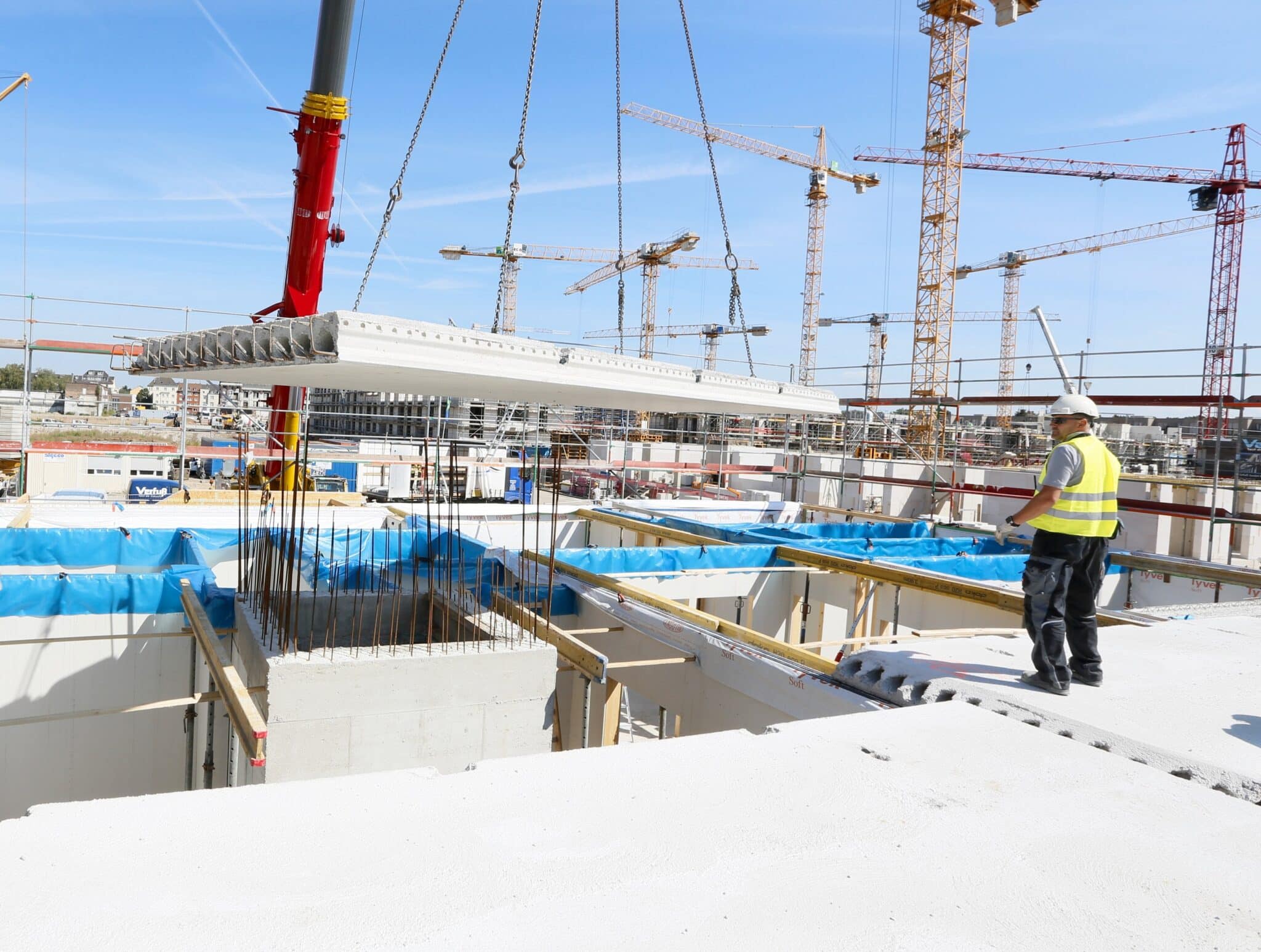 Ein Großkran hebt eine Betonfertigteildecke über einen Rohbau, während ein Bauarbeiter die genaue Positionierung der Deckenelemente überwacht.