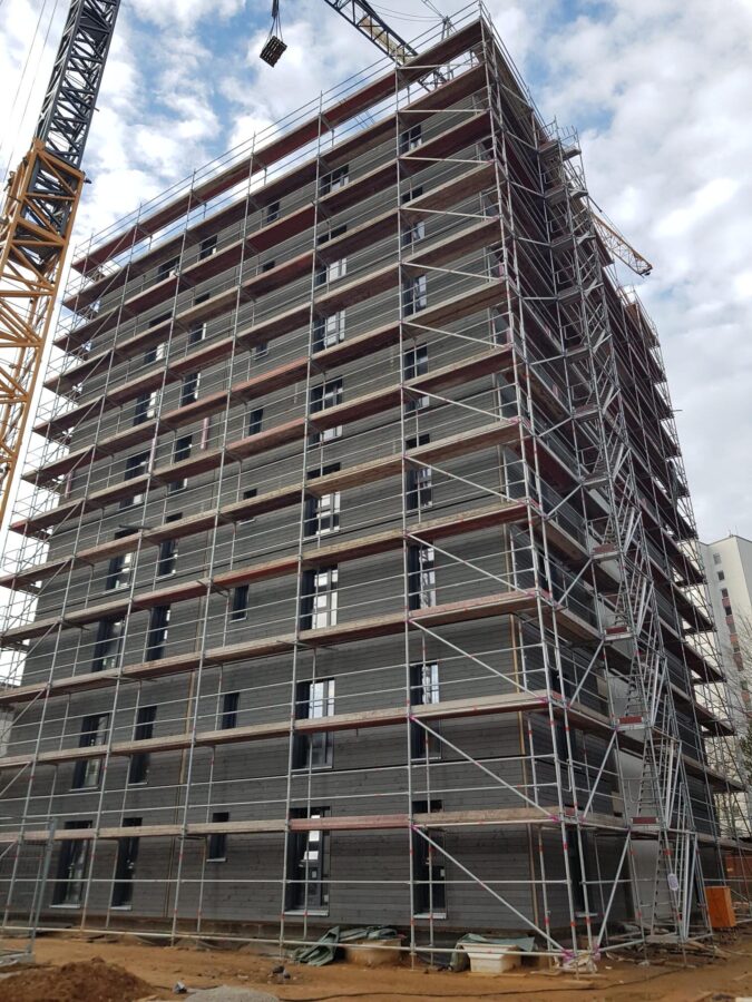 Großformatige Betonfertigteile werden mithilfe eines Turmdrehkrans an einer mehrgeschossigen Gebäudefassade montiert; das gesamte Gebäude ist eingerüstet.