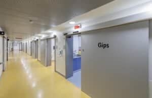 Moderner Krankenhausflur mit gelbem Bodenbelag und grauen automatischen Schiebetüren, eine Tür trägt die Aufschrift „Gips“.