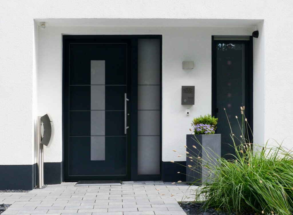 Moderner Hauseingang mit anthrazitfarbener Haustür, seitlichem Glaselement und klar gestaltetem Vorplatz mit Pflanzkübel und Edelstahl-Details.