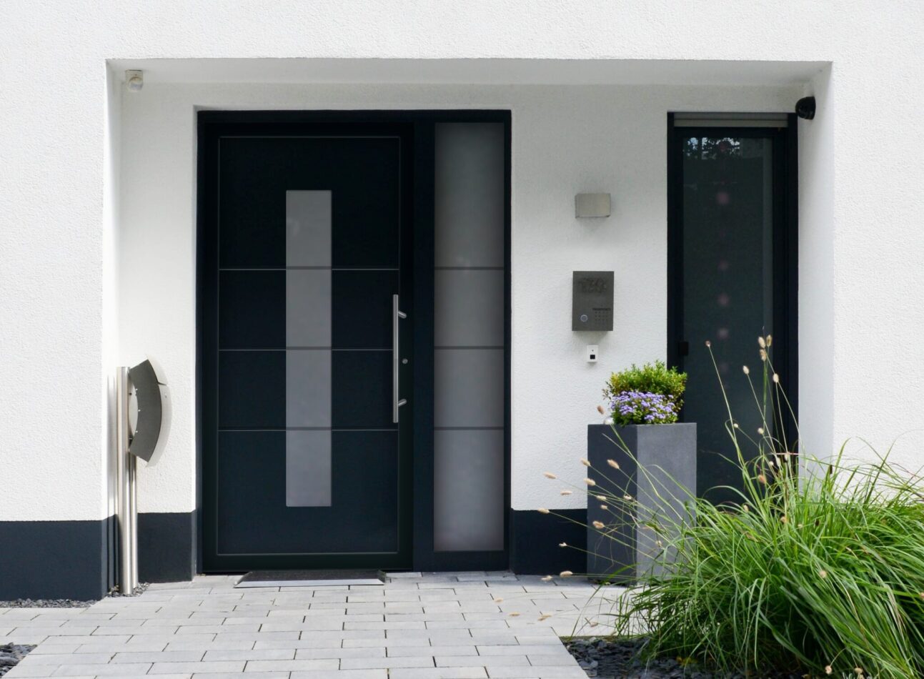 Moderner Hauseingang mit anthrazitfarbener Haustür, seitlichem Glaselement und klar gestaltetem Vorplatz mit Pflanzkübel und Edelstahl-Details.