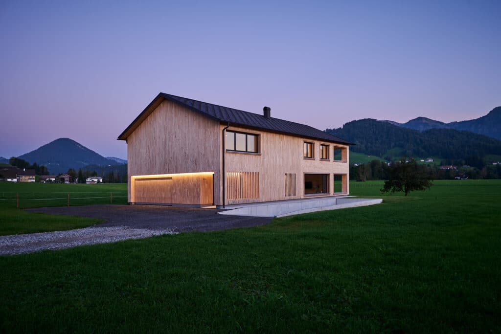 Zweigeschossiges Einfamilienhaus mit Holzfassade und großflächigen Fenstern inmitten einer grünen Wiese, Abendstimmung mit Bergkulisse.